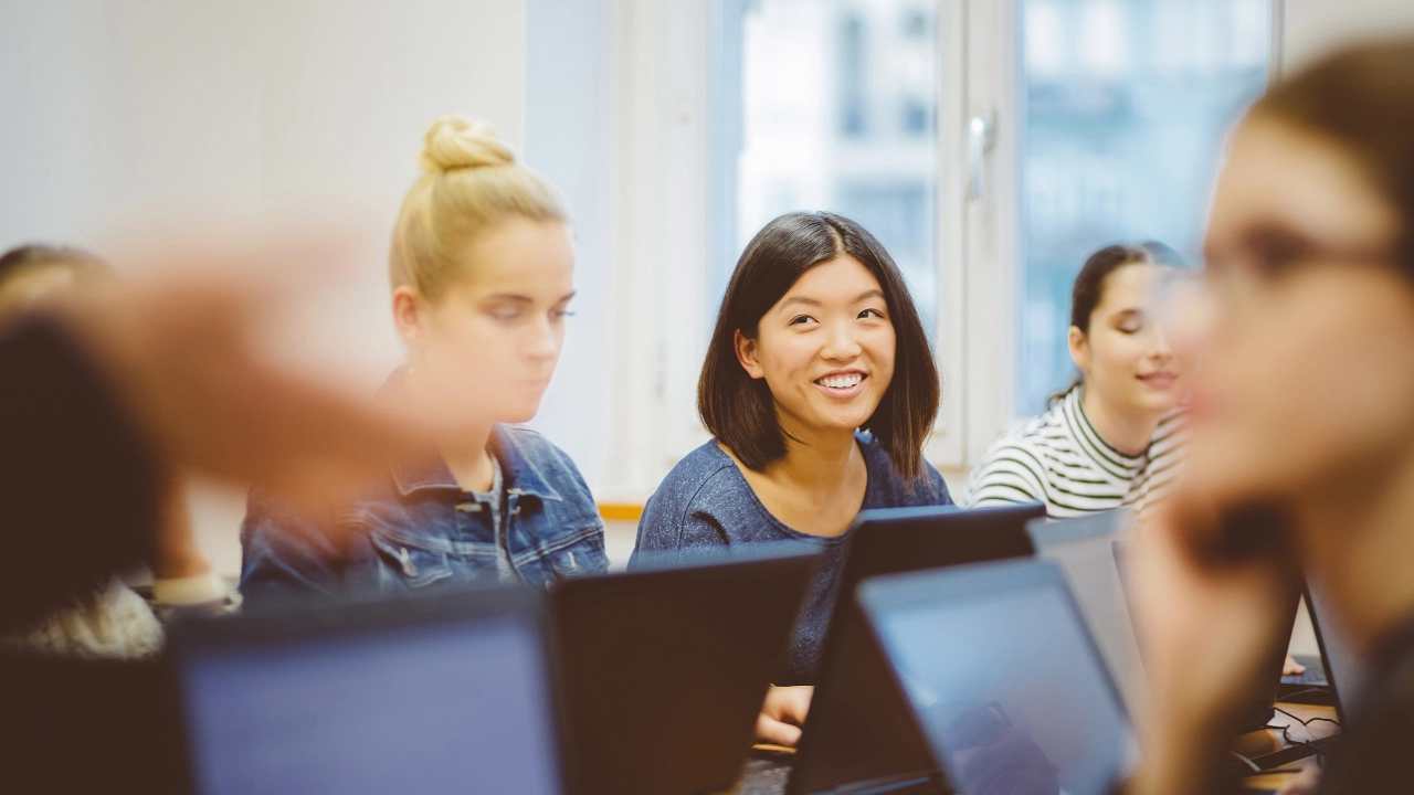 Junge Frau sitzt am Laptop zwischen einigen anderen Seminarteilnehmern und lächelt.
